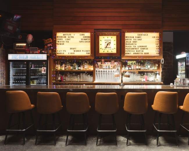 A bar with orange stools, illuminated signage displaying drink options, and shelves lined with glassware is set against a wooden wall backdrop.