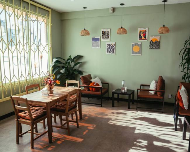 The image shows a cozy café interior with a wooden table and chairs, surrounded by plants and adorned with wall art, under soft illumination from hanging lights.