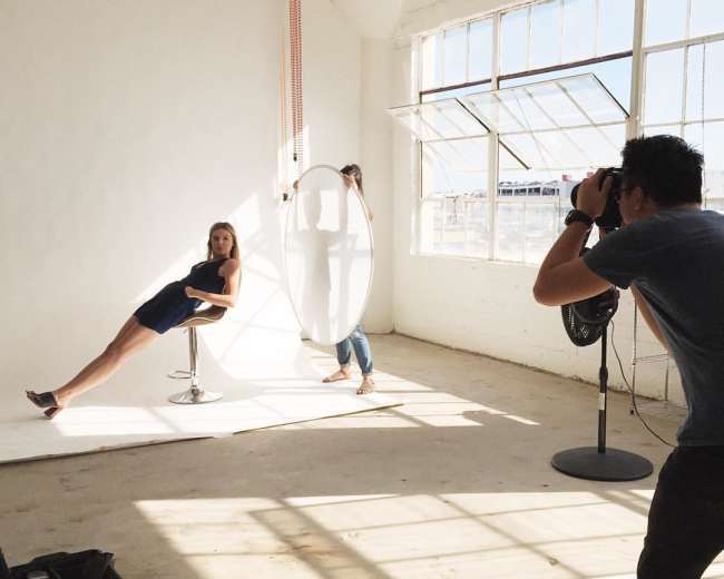 A woman sits on a stool in a well-lit studio while a photographer captures her image and an assistant holds a reflector nearby.