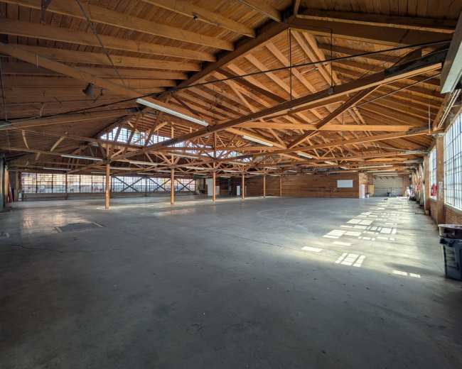 The image shows a large, empty warehouse with a wooden beamed ceiling and concrete flooring, illuminated by natural light from large windows.