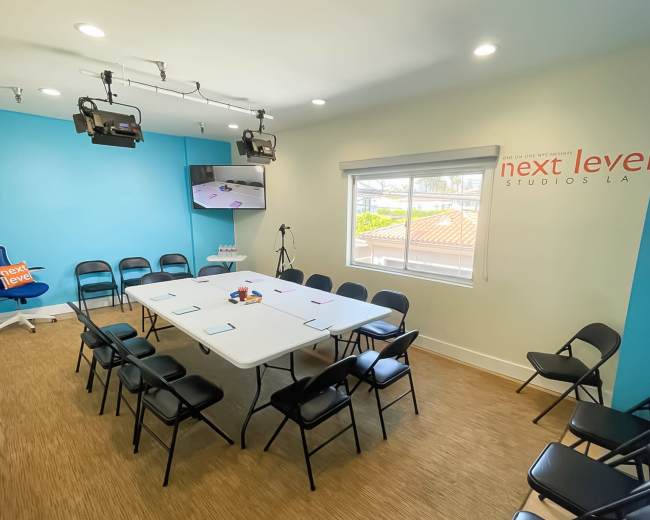 A conference room is arranged with a rectangular table surrounded by black chairs, featuring a screen and lighting fixtures on the ceiling.