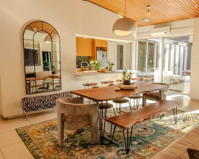 A modern dining area features a wooden table with benches, surrounded by stylish furniture and large glass windows that provide natural light.