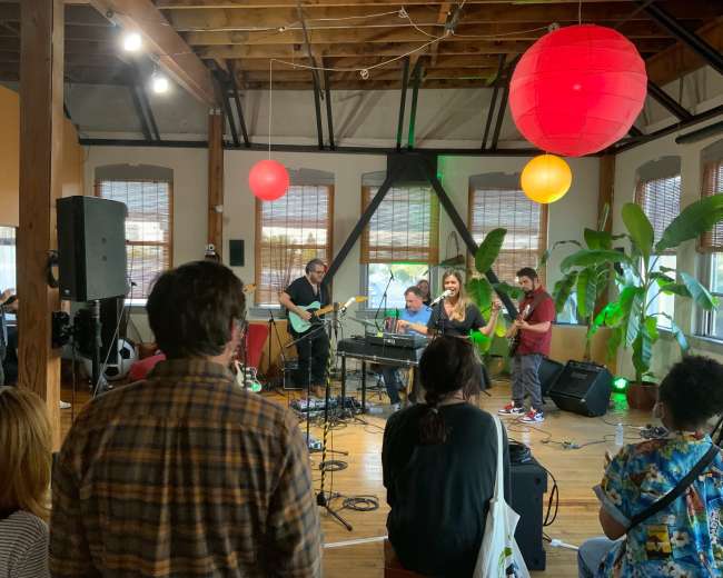 A band performs on stage in a cozy venue with plants and colorful lanterns hanging from the ceiling, while an audience listens attentively.