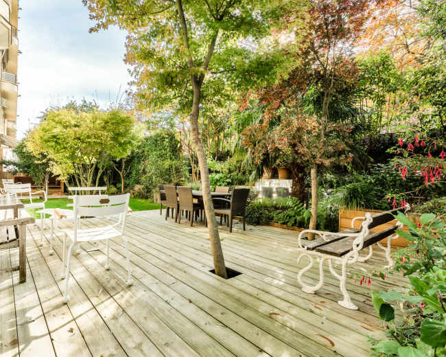 The image shows a wooden deck surrounded by greenery, with several seating areas and a dining table under trees.