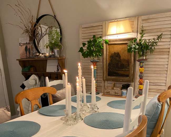 A dining table is set with blue placemats and white candles, surrounded by wooden chairs in a cozy, well-decorated room.