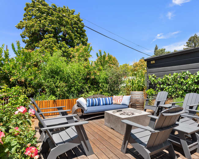 The image shows a wooden deck with a rectangular fire pit surrounded by several gray Adirondack chairs and a blue sofa, set amidst lush greenery.