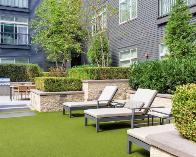 The image shows a landscaped courtyard area with lounge chairs, a barbecue grill, and a dining table amidst greenery and stone features.