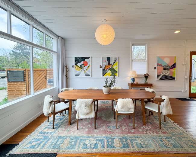 A dining room features a long wooden table surrounded by upholstered chairs on a patterned rug, with large windows and modern artwork on the walls.