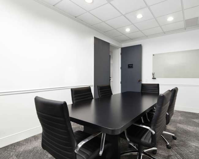 The image shows a modern conference room with a black table and six black chairs, against a white wall with a glass board and a door.