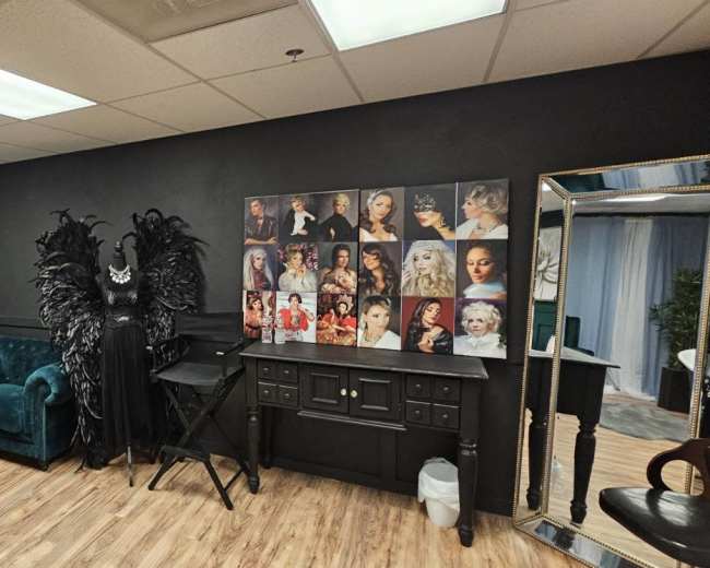 The photo shows a stylish studio space featuring a teal couch, a makeup station, a black wall adorned with portraits, and an ornate black costume with feathers.