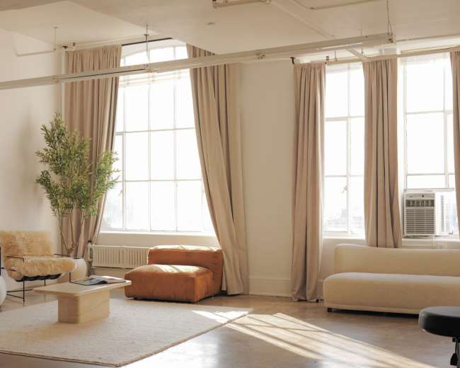 The image shows a bright, minimalist living room with large windows, a plush sofa, an armchair, a coffee table, and potted plants.