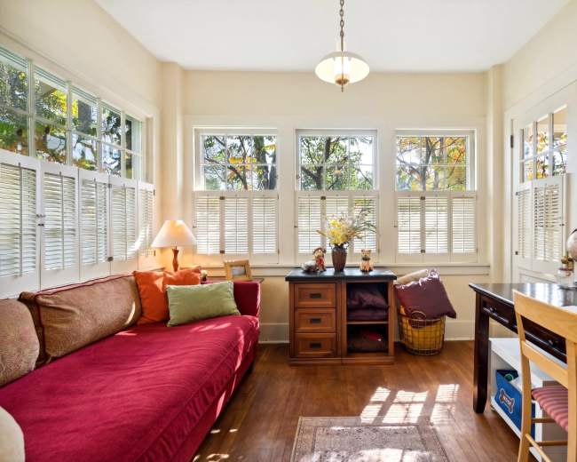 The image shows a cozy sunroom with a red sofa, a wooden desk, and multiple windows letting in natural light.