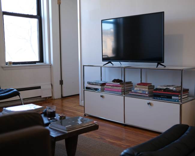 A television sits on a modern stand with open shelves filled with books, next to a black chair and a white wall.