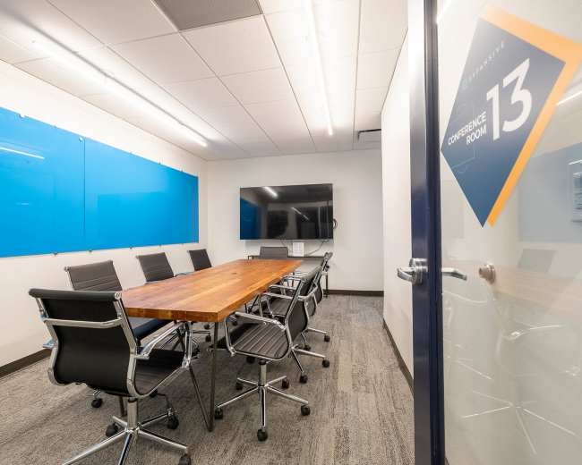 A modern conference room features a long wooden table surrounded by black chairs, a large screen on the wall, and blue presentation boards.