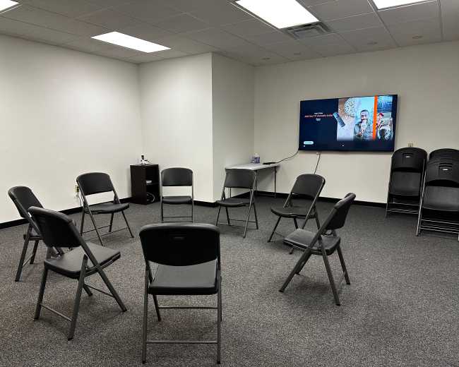 A meeting room contains a circular arrangement of black folding chairs facing a television mounted on the wall.
