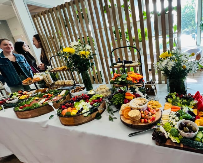 A rectangular table displays an array of colorful food items, including fruits, vegetables, cheeses, and crackers, with floral arrangements in the background.