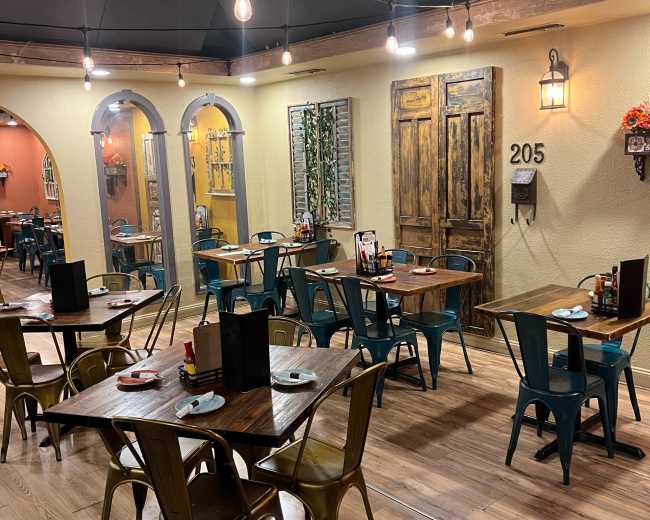 The image shows a restaurant interior with wooden tables, metal chairs, and bright wall colors, featuring decorative elements like framed pictures and light fixtures.