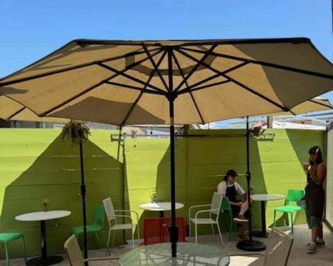 The image shows an outdoor seating area with several tables and chairs under large umbrellas, surrounded by green walls.