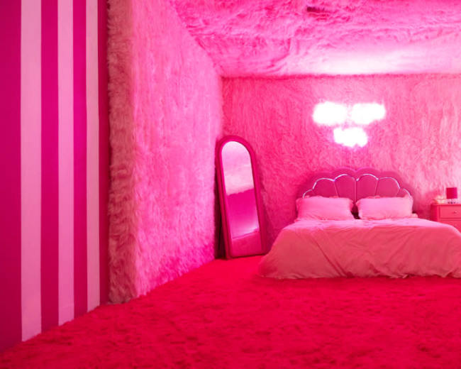 A vibrant pink bedroom with fuzzy walls, a bed with a scalloped headboard, a round mirror, and a nightstand.