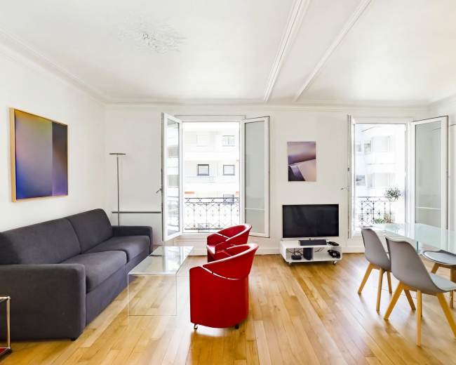 The image features a modern living room with large windows, a gray sofa, red chairs, a glass dining table, and a television against a white wall.