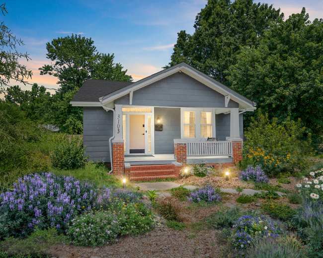The image shows a small gray house with a front porch, surrounded by blooming flowers and lush greenery under a twilight sky.