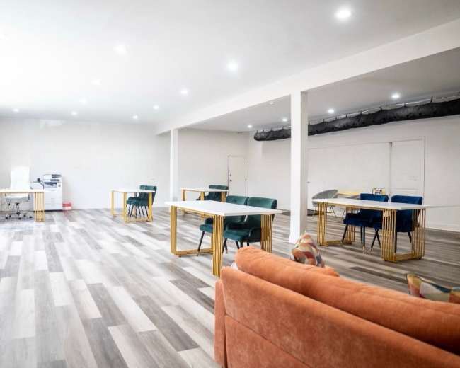 The image shows a spacious, modern office interior featuring several desks, a printer, and a sofa in a neutral-colored room with bright lighting.