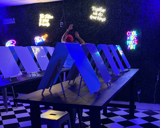 The image shows a brightly lit art studio with several easels set up on tables, surrounded by colorful neon signs.