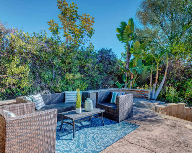 The image shows a outdoor seating area with wicker furniture, a coffee table, and decorative plants in a well-maintained garden.