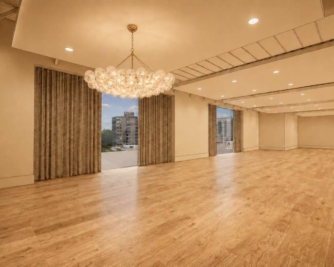 The image shows a spacious interior room with wooden flooring, a chandelier, and large windows that offer a view of an urban landscape.