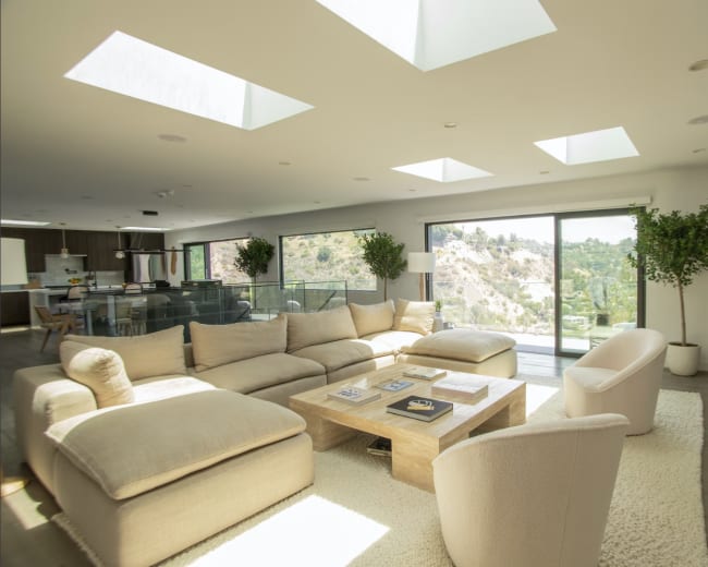 A modern living room features a large, beige sectional sofa, a wooden coffee table, and floor-to-ceiling windows with views of a hillside.