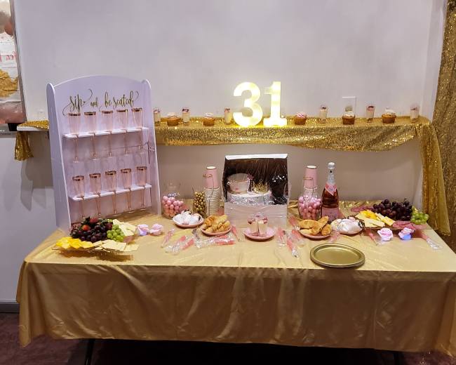 A decorated table featuring a variety of desserts, a cake, and beverages, set against a gold and white backdrop with the number "31" illuminated.