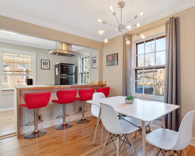 The image shows a modern dining area with a white table and chairs beside a kitchen bar with red stools, illuminated by a contemporary light fixture.