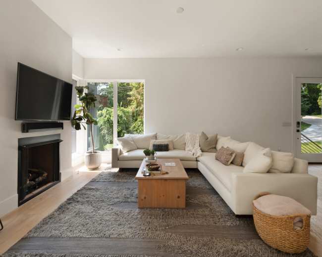 The image shows a bright, modern living room with a large white sectional sofa, a wooden coffee table, a TV mounted on the wall, and large windows allowing natural light.