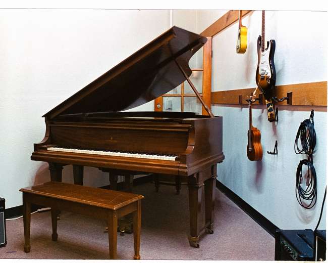 The image features a grand piano positioned in a cozy music room, accompanied by a small bench and various musical instruments hanging on the wall.