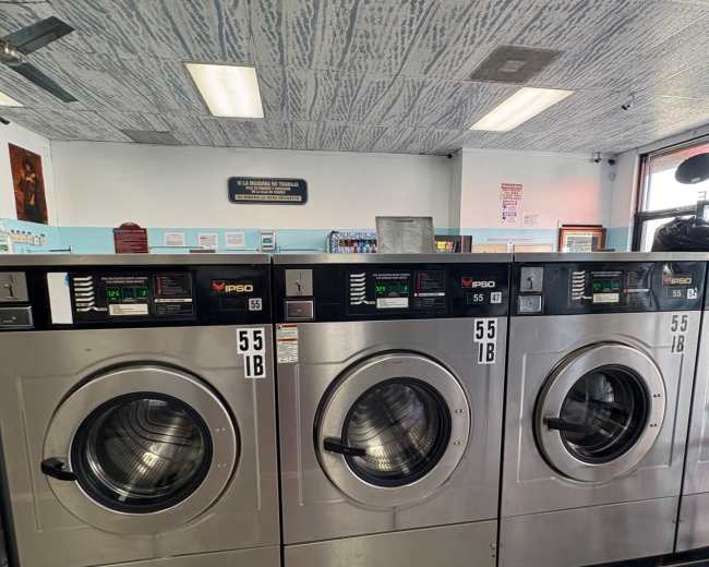 The image shows three large, silver commercial washing machines lined up in a laundromat, featuring digital displays and metallic finishes.