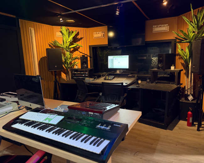 The image depicts a music studio equipped with a mixing console, monitors, speakers, and musical instruments, surrounded by wooden paneling and potted plants.