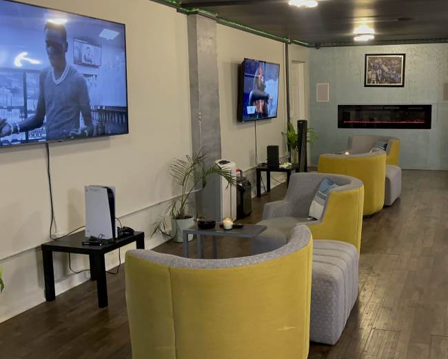 The image shows a lounge area with multiple gray and yellow chairs arranged around low tables, two wall-mounted televisions, and a decorative wall with an electric fireplace.