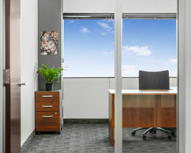 A modern office space features a wooden desk and chair, a filing cabinet with drawers, and a large window with a view of the sky.