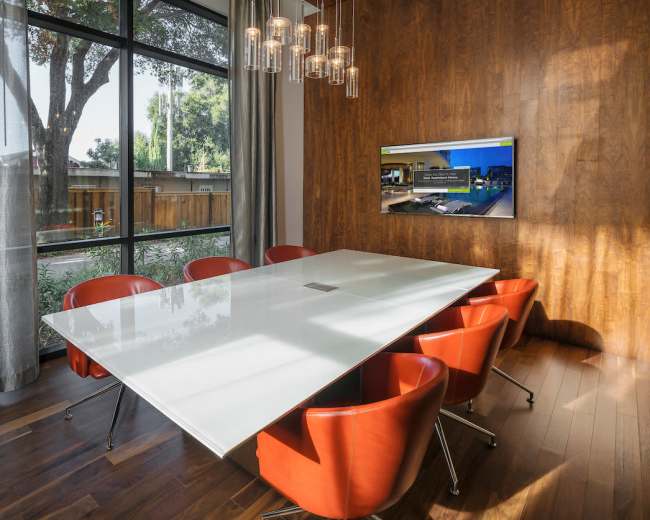 A modern conference room features a white table surrounded by red chairs, large windows with a view of outdoor greenery, and a screen displaying a presentation.