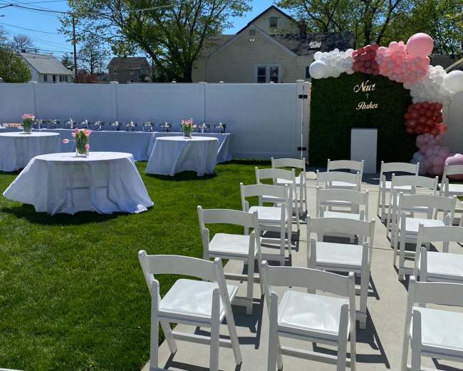 A lawn is set up with white chairs arranged facing a table decorated with flowers, alongside a wall adorned with balloons for a celebration.