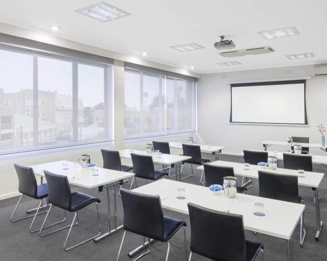 A modern conference room is set up with rows of tables and chairs, featuring a projector screen at the front and large windows allowing natural light to enter.