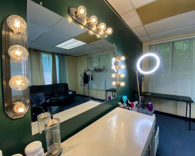 A vanity area is reflected in a large mirror, surrounded by bright lights, with a seating area and clothing rack visible in the background.