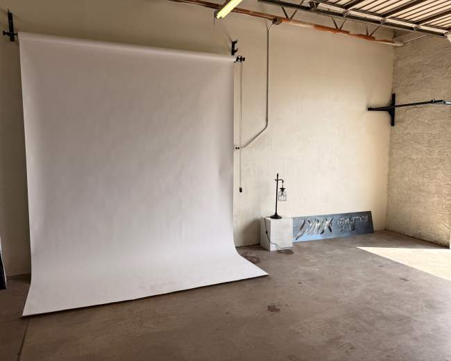 The image shows a photography studio with a white backdrop, a floor lamp, and a metallic sign against the wall.