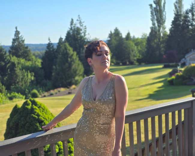 A person in a gold, sequined dress stands on a wooden deck overlooking a landscaped yard with trees and hills in the background.