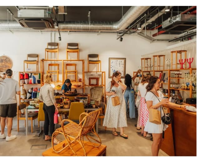A busy retail space features wooden furniture and various artisanal products, with several customers browsing and interacting inside the store.