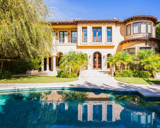 The image shows a large, two-story house with arched windows and a swimming pool in the foreground, surrounded by palm trees and well-maintained landscaping.