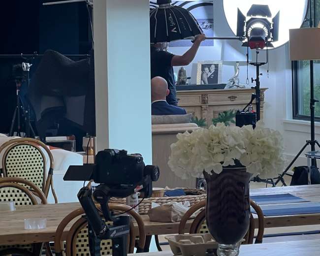 A photography setup is visible in a well-lit room, featuring a photographer with lights and equipment directed toward a subject near a decorative piece.