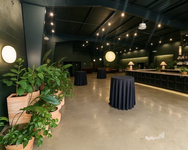 The image shows a spacious event venue with a modern bar, elegant lighting, and potted plants along the walls.