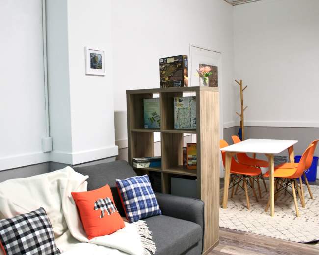 The image depicts a cozy room featuring a gray sofa with colorful pillows, a wooden bookshelf, and a dining table surrounded by orange chairs.