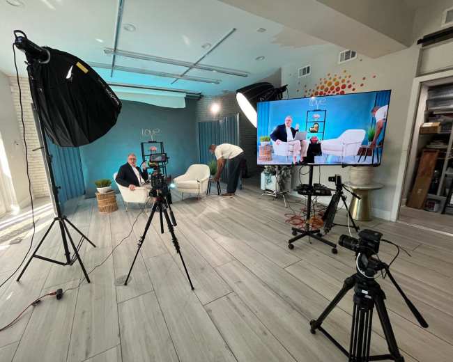 The image shows a video production studio with multiple cameras, lighting equipment, and a monitor displaying a man sitting on a couch against a blue backdrop.